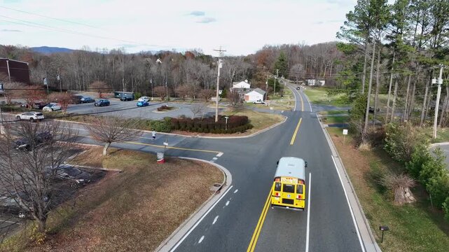 Aerial tracking of shot of yellow school bus making turn on quiet America fall roadway, surrounded by bare trees, small homes, parking lot and distant blue mountains under clear afternoon light.