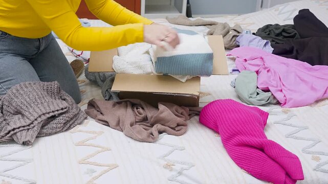 A young woman sorting clothes and packing into cardboard boxes. Donations for charity, help low income families, declutter home, sell online, moving moving into new home or recycling