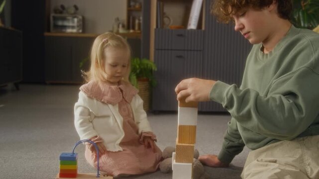 Front view shot of cute little girl sitting on floor at home and playing with wooden toys with older brother building tall tower stacking blocks, copy space