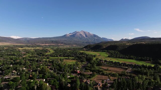 Drone descennd establishing Carbondale patchwork of fields, suburb trees, and golf course, rooftops beneath clear skies and Mount Sopris