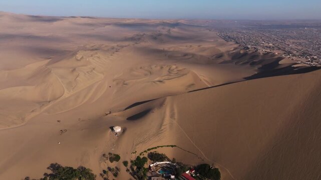 High aerial pullback shows desert ridges surrounding the oasis with long shadows shaping the sandy terrain, establishing Huacachina Peru