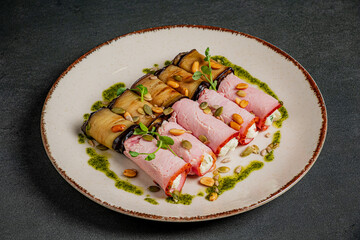 Slices of eggplant and ham are artistically rolled, surrounded by green sauce and seeds