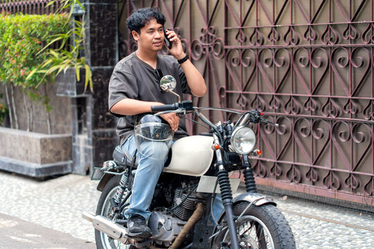 Asian Indonesian Man Using Mobile Phone While Sitting on Motorcycle in Street - Powered by Adobe