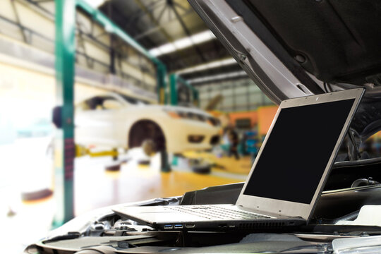 Fototapeta Digital Car Diagnostics, A laptop with blank screen sits on a car engine in a modern auto repair shop, displaying diagnostic data while a vehicle is elevated on a lift for professional service.