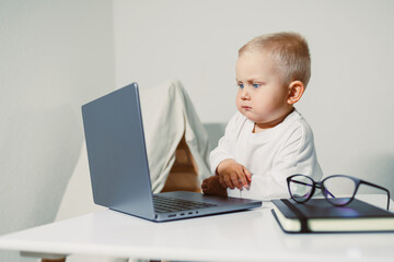 Focused toddler explores the wonders of technology while sitting at a desk with a laptop and glasses nearby in a bright, modern room