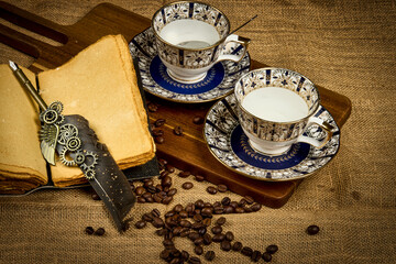 Vintage still life with porcelain coffee cups, coffee beans, antique book and decorative pen on rustic wooden background.