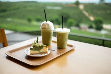Matcha cheesecake served with iced matcha drinks on a wooden tray, placed by a window overlooking lush green hills in a peaceful cafe setting