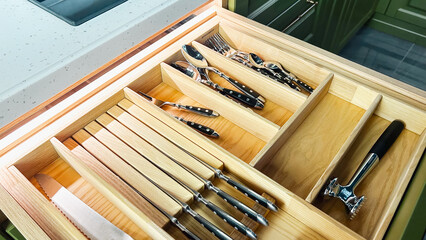 Bright clean kitchen wooden drawer neatly organized with various shiny metal cutlery set and a tenderizer.