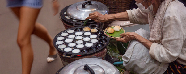 elderly Thai woman vendor prepares authentic Kanom Krok (coconut pancakes) on a hot griddle at a busy street food market, showcasing traditional Asian cuisine and culture