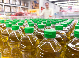 low-angle shot of many clear plastic bottles of golden cooking oil with green caps lined up on a grocery store shelf, with a male customer blurred in the background, symbolizing healthy eating 