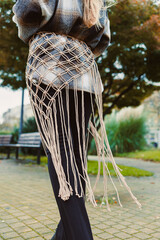 Stylish woman showcases intricate macrame belt while strolling through a park in autumn, surrounded by vibrant trees and unique urban scenery