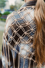 Bright strands of hair flow down a woman's back as she showcases a beautifully crafted macrame shawl in a vibrant outdoor setting near a park