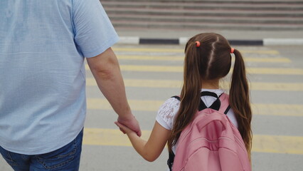 father’s care on a busy street, Child in ponytails heading to school, Safety at pedestrian crossing, Morning rush with family, Holding hands for security, Crossing road carefully, Pink backpack