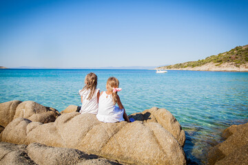 Fototapeta premium Two young girls in white dresses sit on large coastal rocks by a clear blue sea