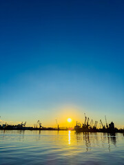 Vibrant sunset scenery over the water with industrial cranes and ships silhouetted against a clear blue sky
