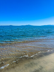 Waves gently lap against the sandy shore under a bright blue sky with distant islands on the horizon during a sunny afternoon at the beach