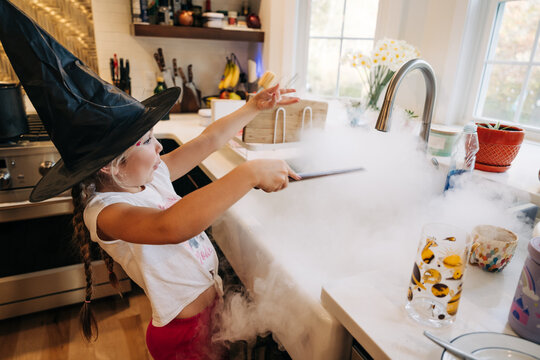 Girl in witch hat plays with foggy potion at kitchen sink
