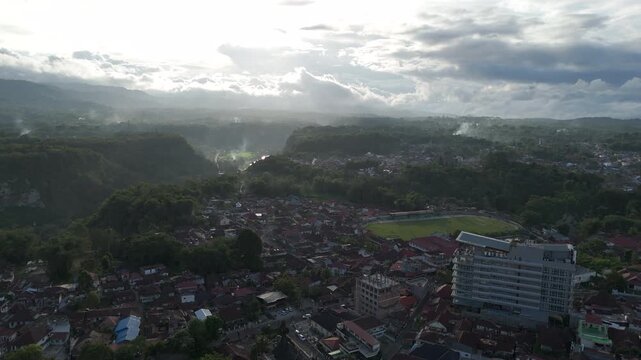 Aerial view of Bukittinggi city, West Sumatra, featuring a major hotel (Santika) and stadium situated on the edge of the lush, fog-filled Sianok Canyon. Dramatic backlight creates a moody atmosphere.