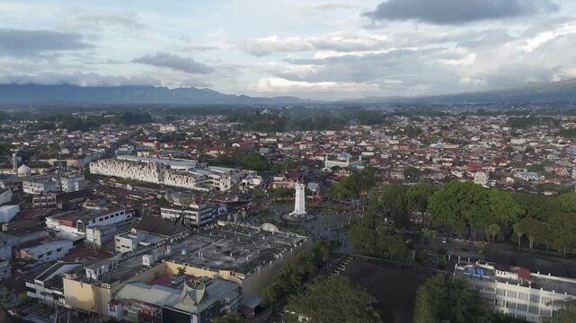 Wide aerial drone shot over the bustling downtown square of Bukittinggi, West Sumatra, featuring the iconic Jam Gadang clock tower monument, market area, and dense surrounding city.