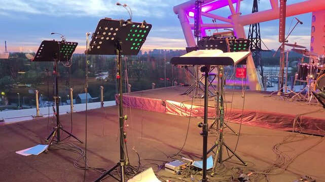 Outdoor concert stage prepared for live performance at sunset, Professional music equipment with stands, cables and city skyline during evening setup