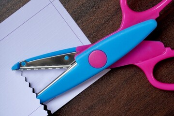 Pink and blue colored zig zag craft tool scissors object with white paper isolated on horizontal brown table surface background.