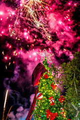 Outdoor winter celebration with decorated Christmas tree, red bows and ornaments glowing under night sky, bright fireworks display above, festive New Year atmosphere and seasonal holiday mood