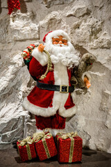 Santa Claus figurine holding teddy bear and gift bag standing near wrapped presents against stone wall, Christmas decoration concept, reminder of holiday gifts, winter festive scene
