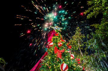 Festive outdoor celebration featuring glowing Christmas tree with red bows and ornaments, colorful fireworks above, New Year event under night sky, winter holiday spirit, cheerful atmosphere