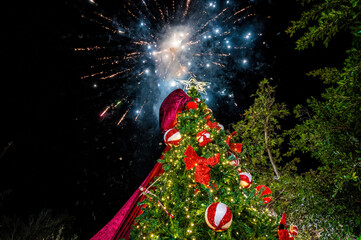 Outdoor holiday scene with illuminated Christmas tree decorated with red bows and ornaments, vibrant fireworks bursting across night sky, New Year celebration, winter festival atmosphere, joyful festi