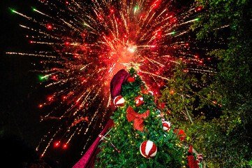 Illuminated Christmas tree with red bows and ornaments standing outdoors as colorful fireworks burst across night sky, creating joyful holiday celebration scene with New Year energy and winter festiva