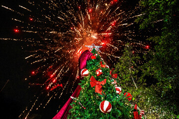 Festive Christmas tree decorated with red bows and ornaments under night sky with bright fireworks explosion above, holiday celebration outdoors, New Year atmosphere, winter festival mood