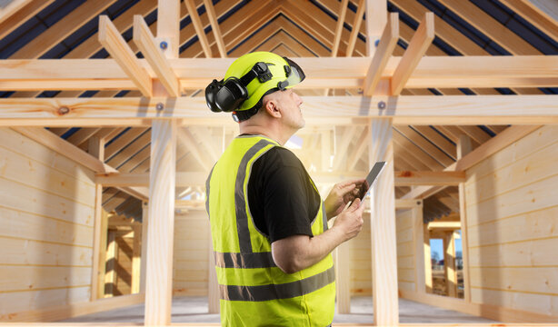 Wooden house construction supervisor reviews structural elements with a tablet, overseeing framework assembly and ensuring quality standards on site.