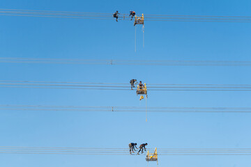 Aerial view of linemen perched on platforms attending to high-voltage power lines against a clear blue canvas, Joypurhat, Rajshahi Division, Bangladesh.