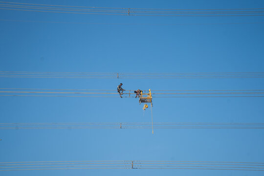 Aerial view of linesmen precariously working on high-tension power lines against a bright blue sky, Joypurhat, Rajshahi Division, Bangladesh.