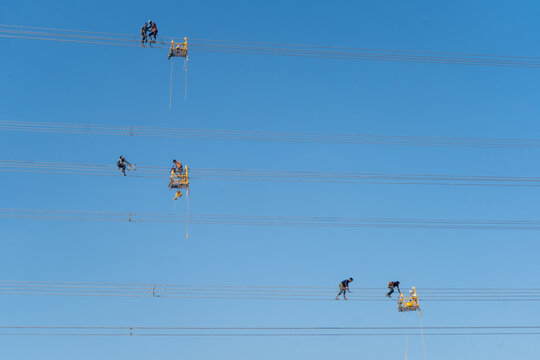 Aerial view of workers perched precariously on high-tension power lines against a stark blue sky, Joypurhat, Rajshahi Division, Bangladesh.
