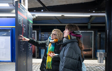 Two Caucasian women in winter clothing checking a subway information board in a metro station, illustrating public transportation, urban navigation and travel planning in a modern European city