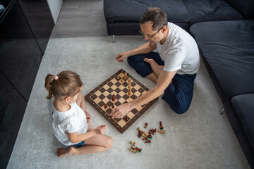 Father and daughter sitting on the floor and playing chess together in the living room. Educational...