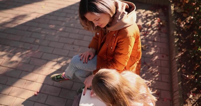 Mother sitting on park parapet helps daughter read school book. Family spends study time together while enjoying fresh air near bushes slow motion