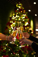 Hands raising glasses of champagne for a festive toast in front of a decorated Christmas tree. Warm holiday atmosphere, bokeh lights, celebration mood and New Year party vibes.