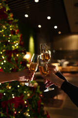 Hands raising glasses of champagne for a festive toast in front of a decorated Christmas tree. Warm holiday atmosphere, bokeh lights, celebration mood and New Year party vibes.