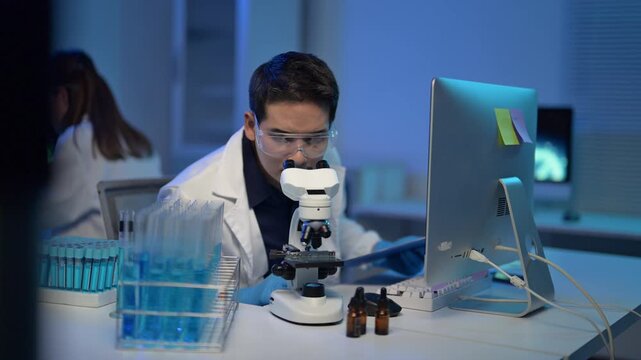A man in a lab coat is working on a computer. There are several bottles on the table, and a microscope is nearby