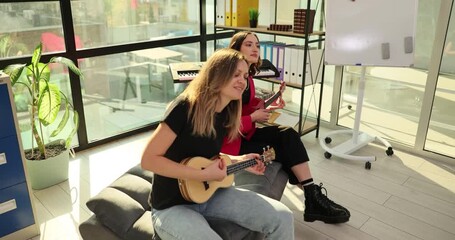 Women strum guitars and sing with lively enthusiasm. Females sit on sofa in bright corporate lounge and perform playful melodies. Slow motion