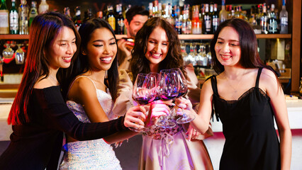 Diverse group of four women, Asian and Caucasian, smiling and clinking wine glasses in a vibrant bar or party setting, capturing joy, celebration, unity, nightlife, friendship, and social bonding.