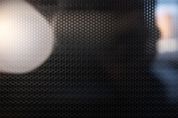 Macro shot of a black perforated industrial grille. The textured surface creates a geometric pattern with blurred lights and reflections for an abstract effect.