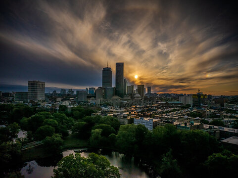Aerial view of a golden sunset casting long shadows over the John Hancock Tower and Prudential Center, illuminating the lush green canopy below, Boston, Massachusetts, United States.