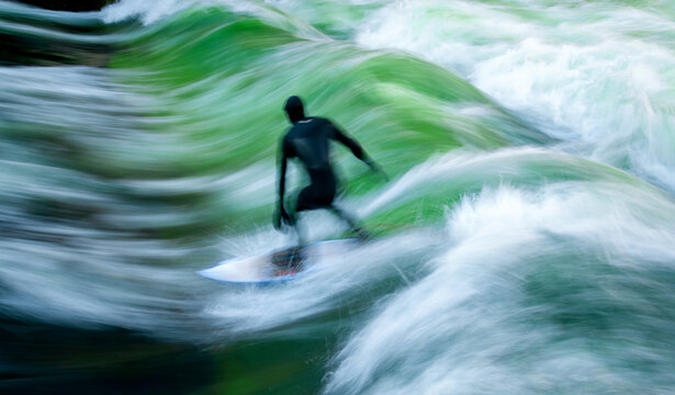 Silhouette of a surfer in a dark wetsuit surfing a standing wave on the Eisbach in Munich (Bavaria). Motion blur caused by long exposure and the flow of the water. Outdoor sport in a park in Germany.