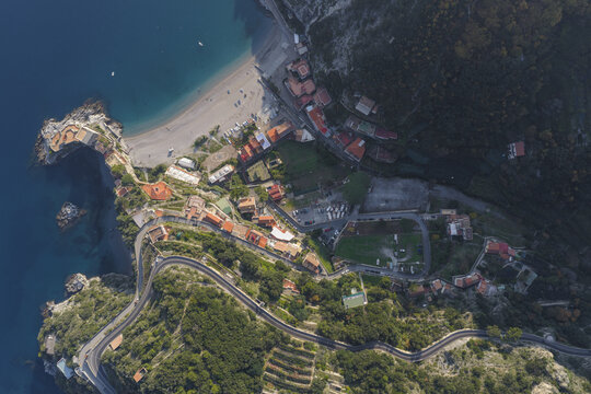 Aerial view of a winding coastal road leading to a sandy beach and the village, nestled between the turquoise sea and the dark green hillside, Erchie, Amalfi Coast, Campania, Italy.