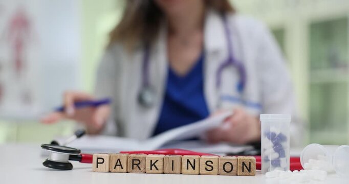 Wooden blocks spell word Parkinson in front of stethoscope and papers. Woman doctor writes neurology report discussing movement disorder risks