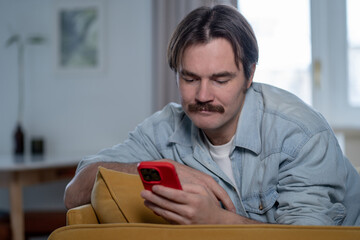 Happy man lounging on couch at home, scrolling through phone with joy. Relaxed interested young male smiling reading something pleasant on smartphone resting comfortably in apartment. Online shopping.