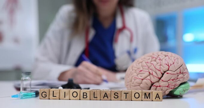 Wooden blocks spell word Glioblastoma in front of brain model on table. Woman doctor writes oncology report studying aggressive brain tumor case